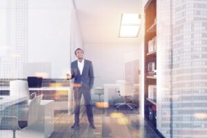 גבר בחליפה מחזיק כוס, עומד ומחייך במשרד מודרני עם חלונות גדולים, שולחנות ומדפים, ומבנים עירוניים נראים מבחוץ.