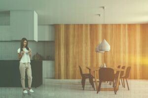 אישה עומדת במטבח מודרני, מביטה בטלפון שלה ומחזיקה כוס. בחדר יש לוחות עץ על הקירות, שולחן אוכל עם שישה כיסאות ותאורה תלויה, ואור השמש חודר פנימה.