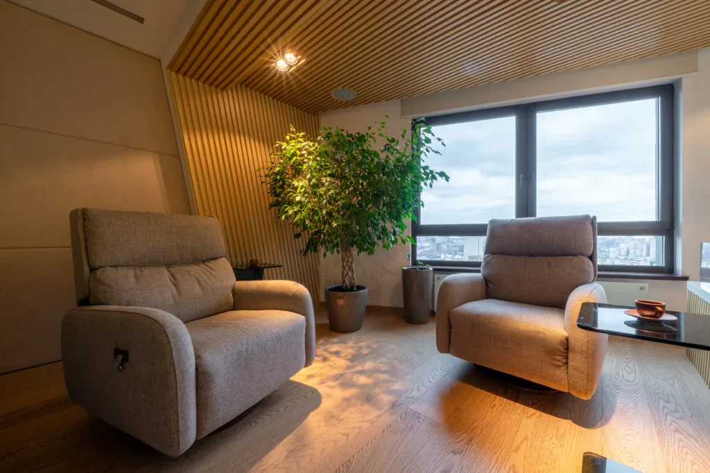 שני כורסאות אפורות עומדות זו מול זו בחדר מודרני עם רצפות וקירות מעץ, חלון גדול המשקיף על העיר, עציץ גבוה ושולחן צד עם כוס ותחתית.