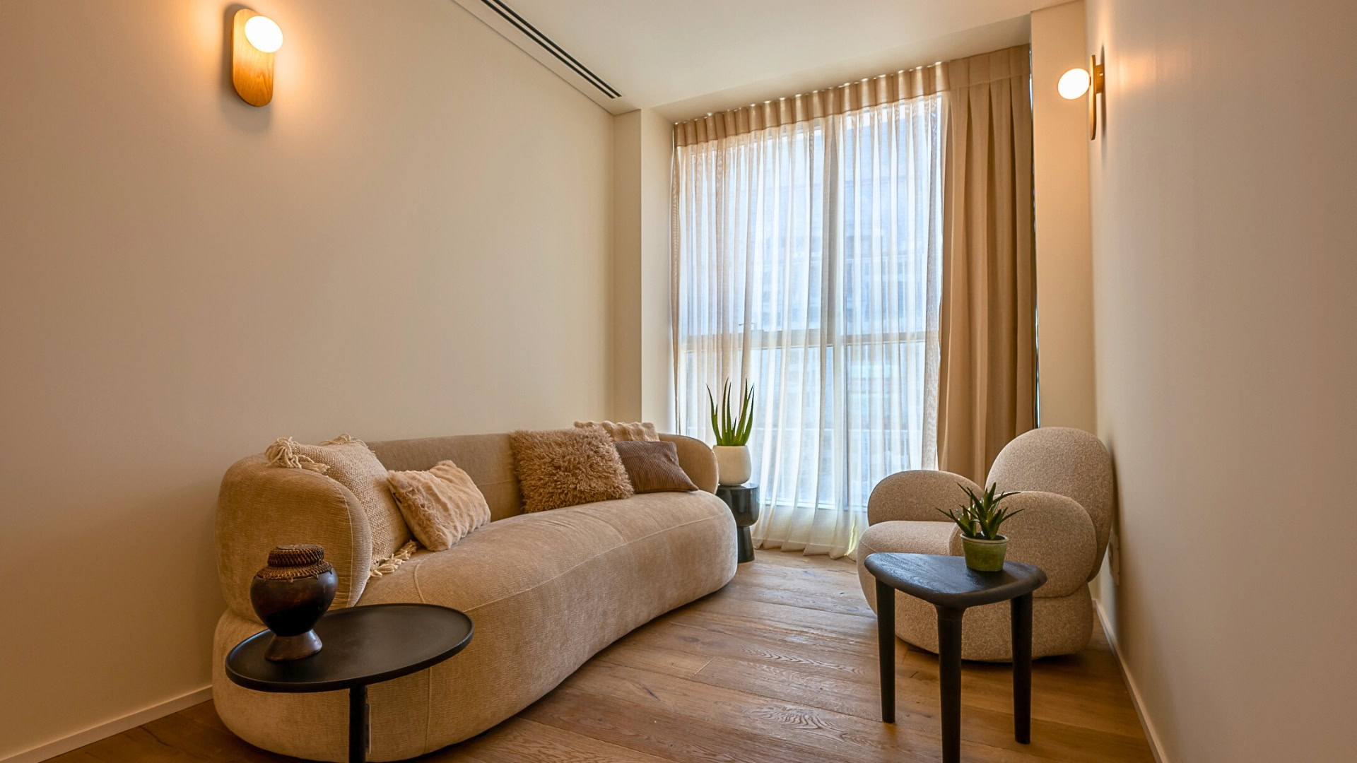 סלון מודרני עם ספה מעוגלת בצבע בז', כיסא עגול בצבע בז', שני שולחנות צד קטנים בצבע שחור עם עציצים ואגרטל, רצפת עץ וחלונות גדולים עם וילונות שקופים המאפשרים כניסת אור טבעי.