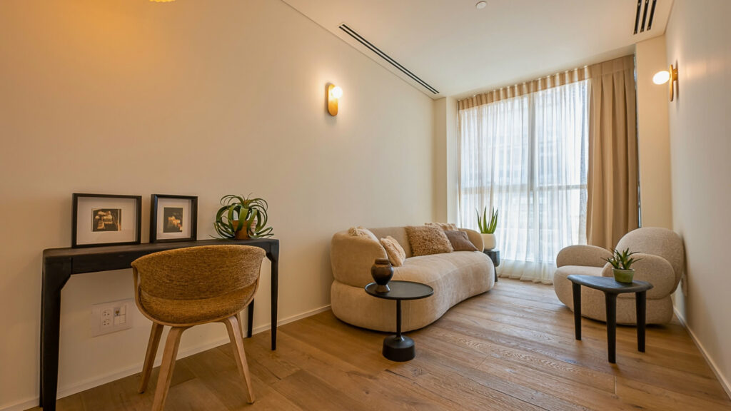 סלון מודרני עם ריצוף עץ בהיר, ספה בצבע בז', כורסה תואמת, שני שולחנות עגולים קטנים עם צמחים, שולחן עבודה שחור עם תמונות ממוסגרות, כיסא קלוע וחלונות גדולים עם וילונות שקופים.