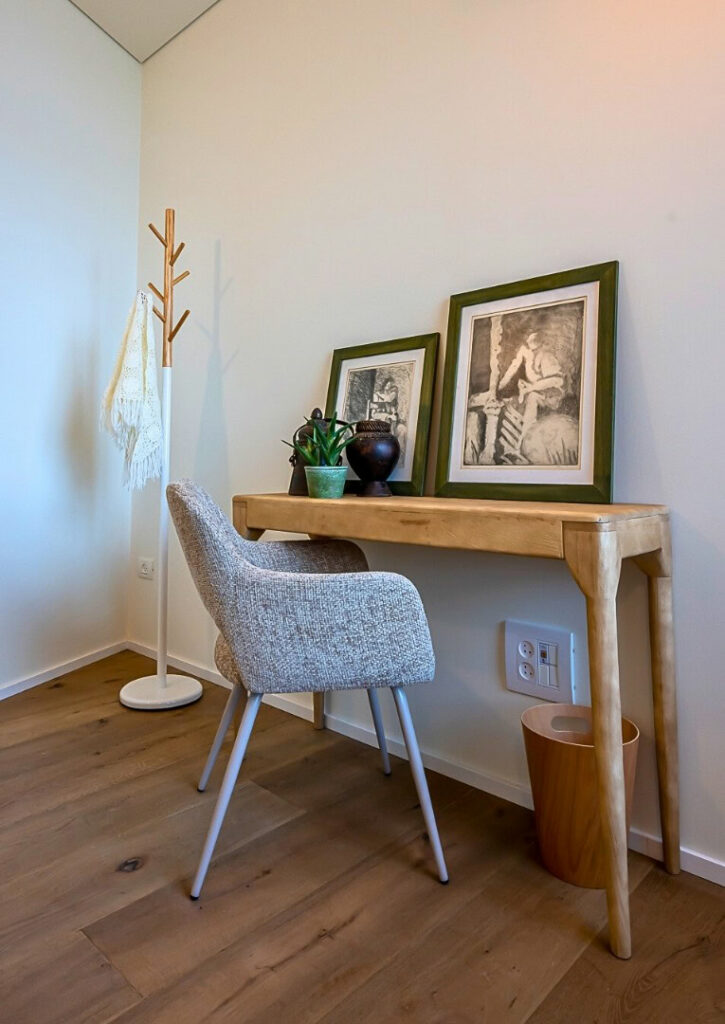 שולחן עץ קטן עם שתי יצירות אמנות ממוסגרות, עציצים וכיסא אפור ניצב בפינה. מתלה מעילים עם צעיף לבן וסל אשפה מעץ נמצאים בסמוך, על רצפת עץ בהירה.