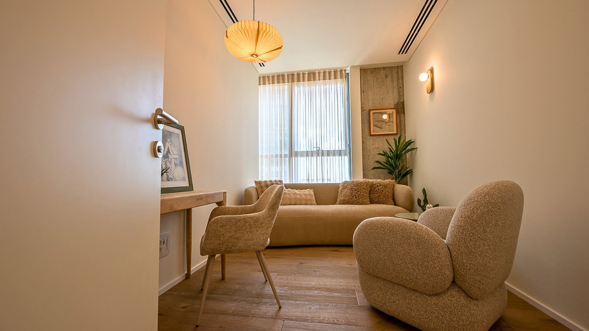 סלון נעים ומודרני עם ספה בצבע בז', כורסה מפנקת, שולחן וכסא מעץ, עציץ, תמונות קיר ותאורה רכה, עם חלון גדול ווילונות שקופים המאפשרים כניסת אור טבעי.