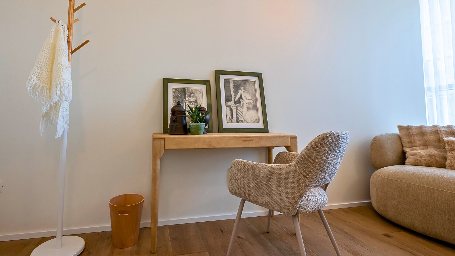 פינת משרד ביתית נעימה עם שולחן עץ, שתי יצירות אמנות ממוסגרות, עציצים, כיסא בז' מרקם, מתלה מעילים עם צעיף לבן, פח אשפה קטן וספה בז' עם שמיכה.