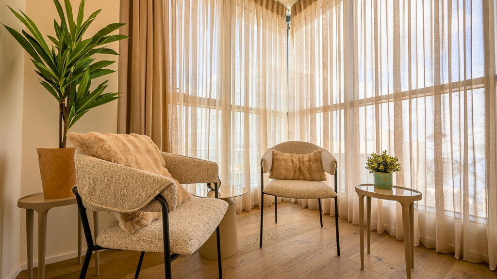 שני כיסאות נוחים עם כריות מסודרים סביב שולחן עגול קטן ועליו עציץ. אור זורם מבעד לווילונות שקופים מהרצפה עד התקרה, ויוצר פינה חמימה ומזמינה. צמח ירוק גבוה ניצב בסמוך.