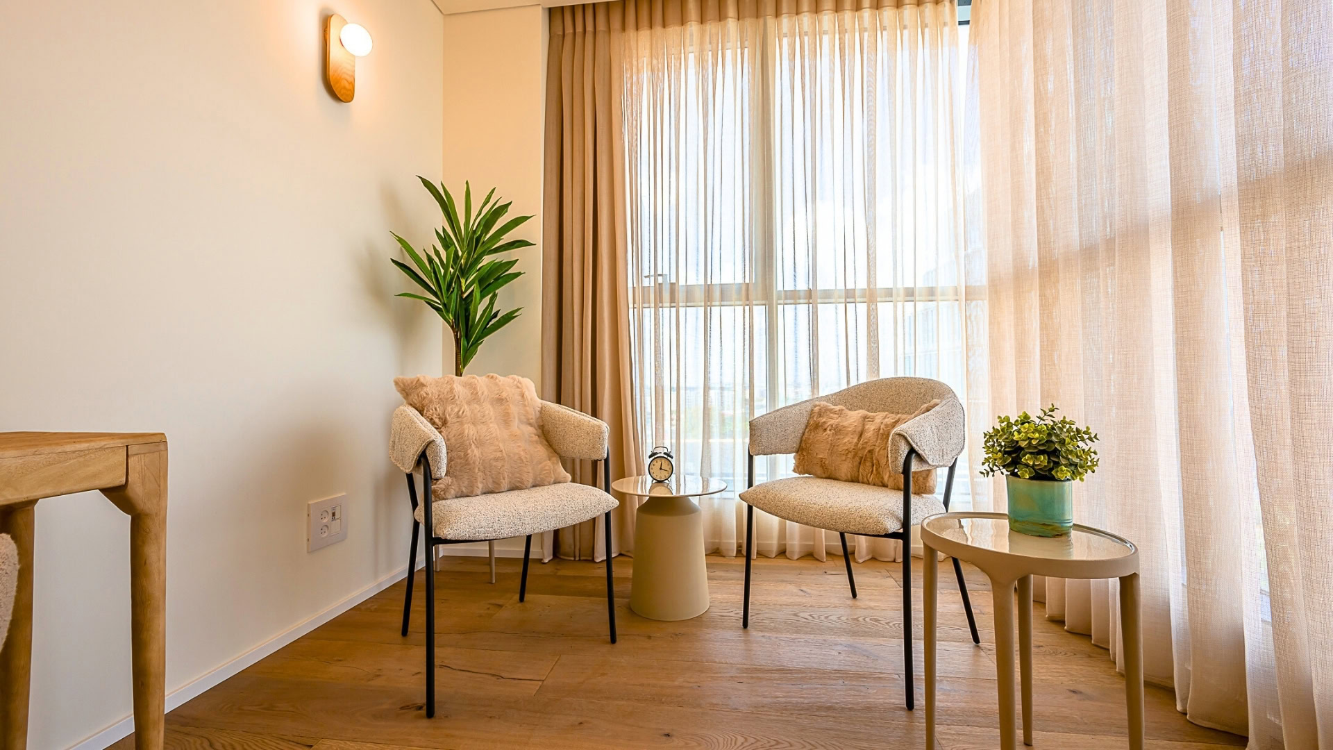 שני כיסאות בצבע בז' עם כריות מונחים זה מול זה ליד שולחן עגול קטן ועליו שעון מעורר. בפינה עומד עציץ, וחלונות גדולים עם וילונות שקופים מכניסים אור טבעי. עציץ נוסף מונח על שולחן אחר.