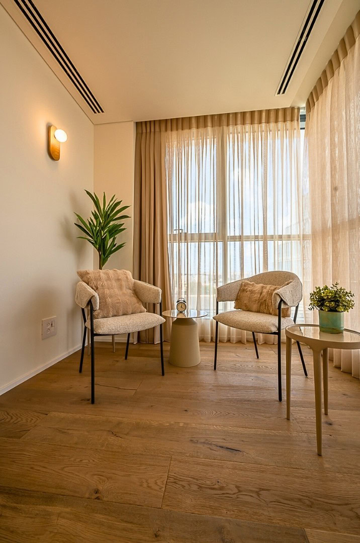 פינת ישיבה נעימה ומוארת עם שני כיסאות מרופדים, שולחן עגול קטן עם עציץ, שולחן צד עם שעון, עציץ, רצפת עץ ווילונות בז' שקופים המכסים חלונות גבוהים.