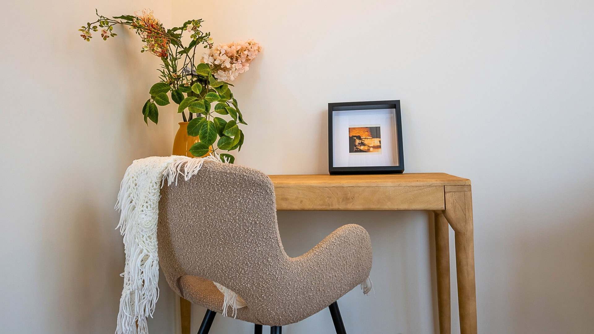 כיסא בז' נעים עם שמיכת סריג לבנה מונחת עליו ניצב מול שולחן עץ. על השולחן עומדים אגרטל עם פרחים ירוקים וכתומים ותמונה קטנה ממוסגרת על רקע קיר בהיר וחלק.