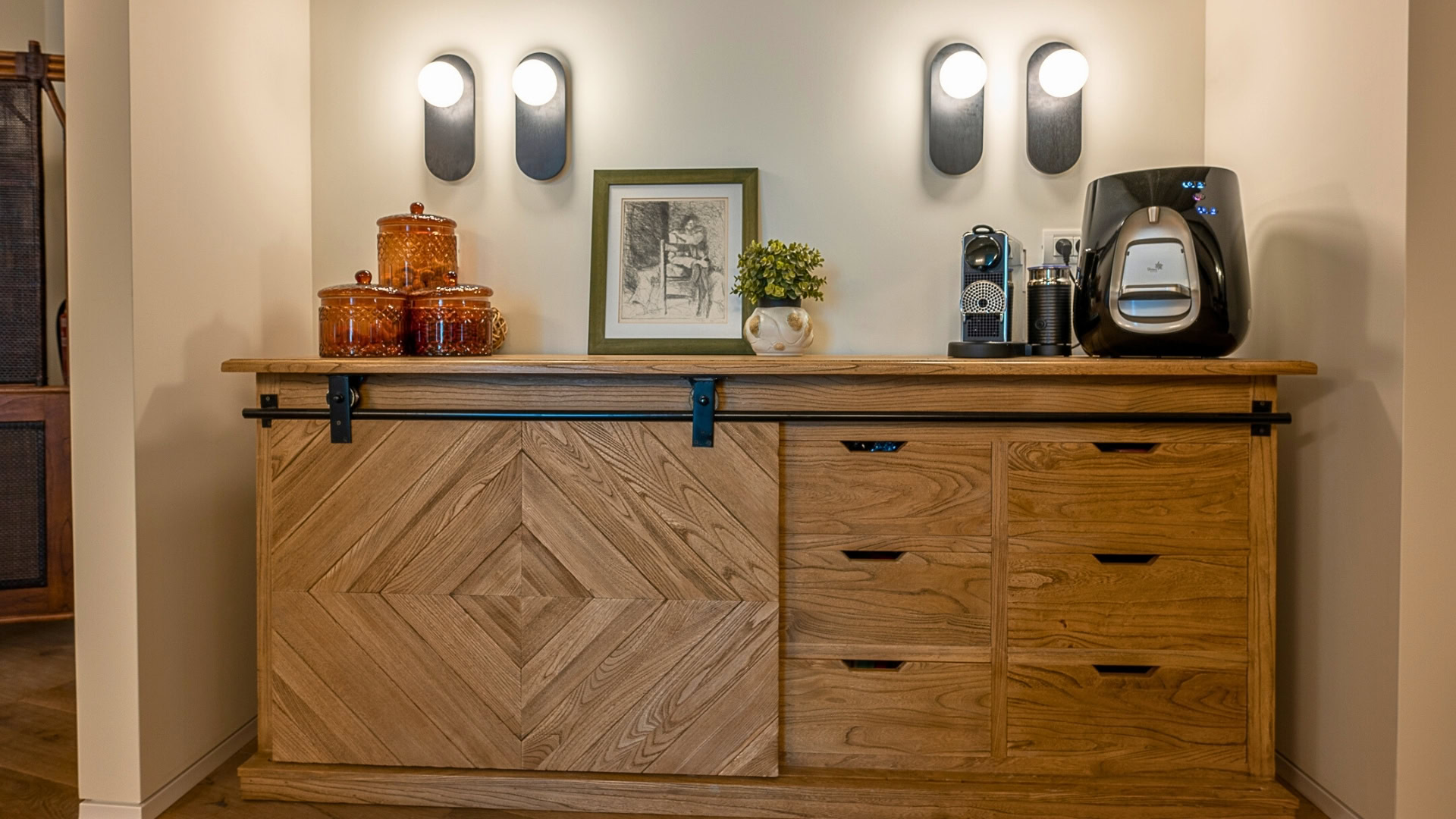 ארון עץ עם דלתות הזזה מכיל צנצנות זכוכית, תמונה ממוסגרת, צמח קטן, מכונת קפה ומכשיר להכנת קפה. שלוש מנורות קיר מותקנות מעל הארון ומאירות את החלל.