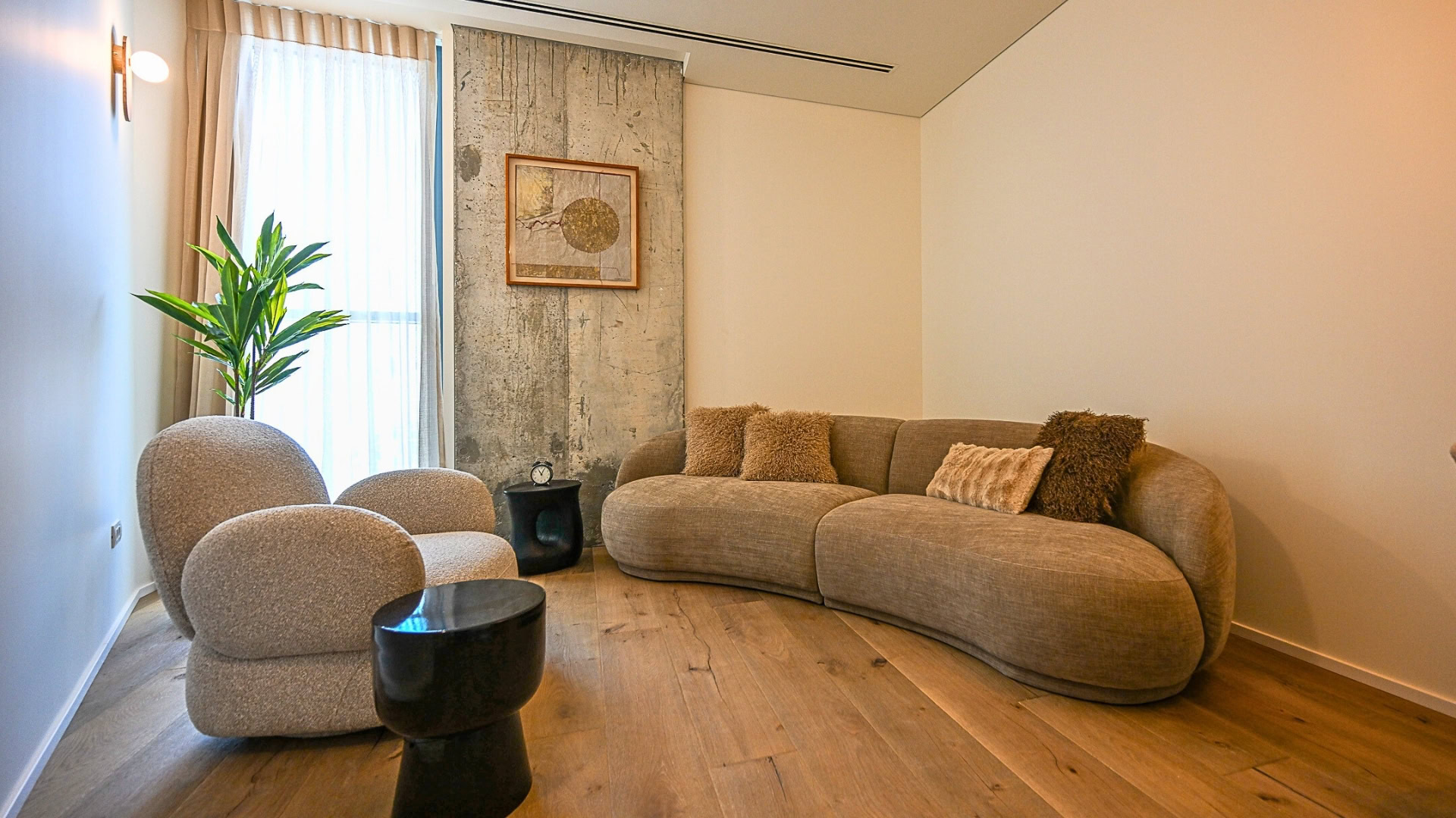 סלון מודרני עם ספה בצבע בז' מעוקלת, כורסה מעוגלת, שולחן צד קטן בצבע שחור, עציץ, ריצוף עץ, קיר עם יצירת אמנות מופשטת וחלונות גבוהים עם וילונות שקופים.