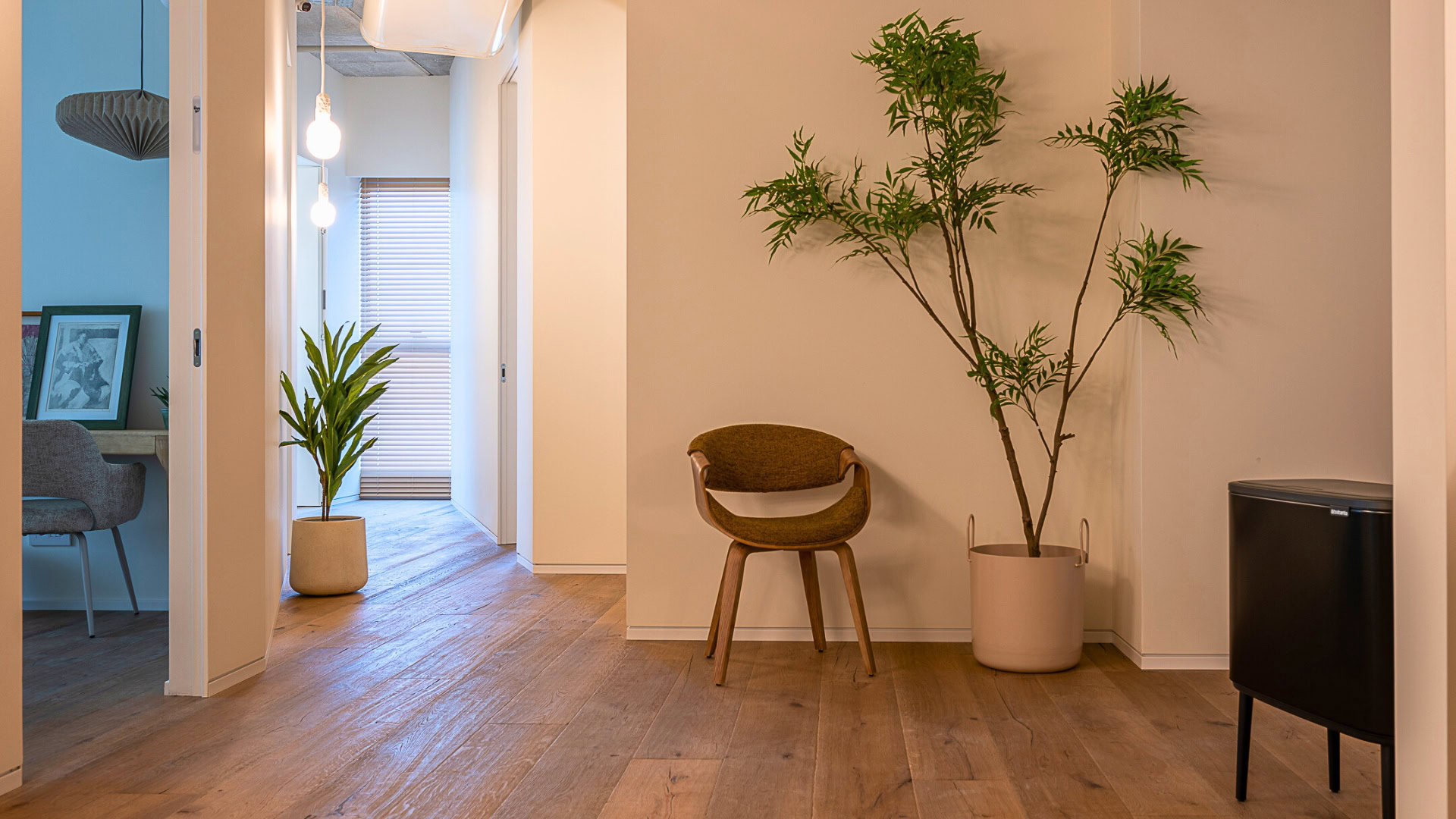 מסדרון מודרני עם ריצוף עץ, כיסא עץ ועציץ גבוה. מנורות תלייה תלויות מהתקרה, וניתן להבחין בפינת עבודה מבעד לדלת פתוחה.