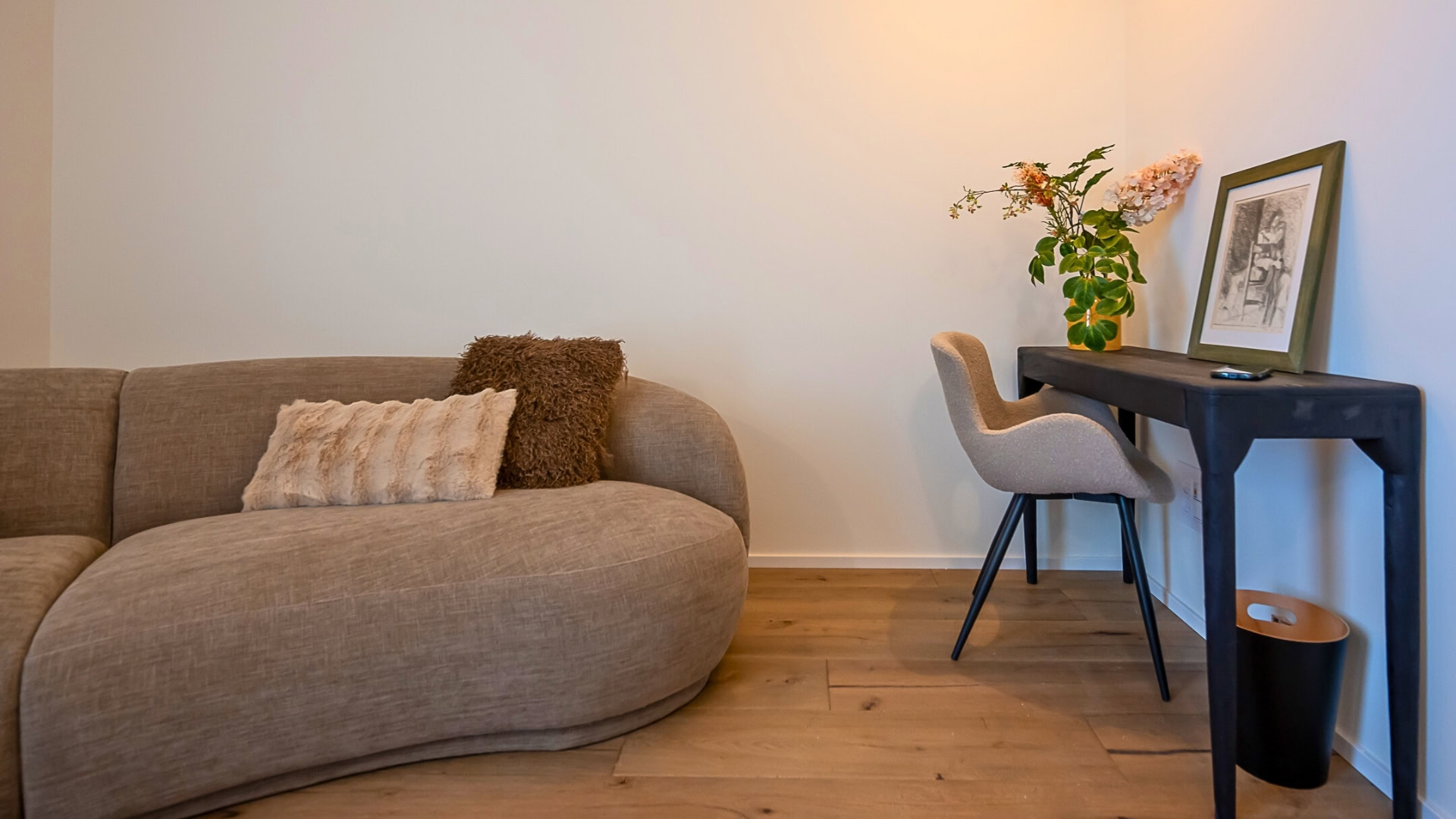 חלל מגורים מודרני עם ספה בצבע בז' מעוקלת, שתי כריות מרקמות, שולחן עבודה קטן בצבע שחור עם כיסא בצבע בז', צמח, יצירת אמנות ממוסגרת ושלט רחוק, כולם מונחים על רצפת עץ.