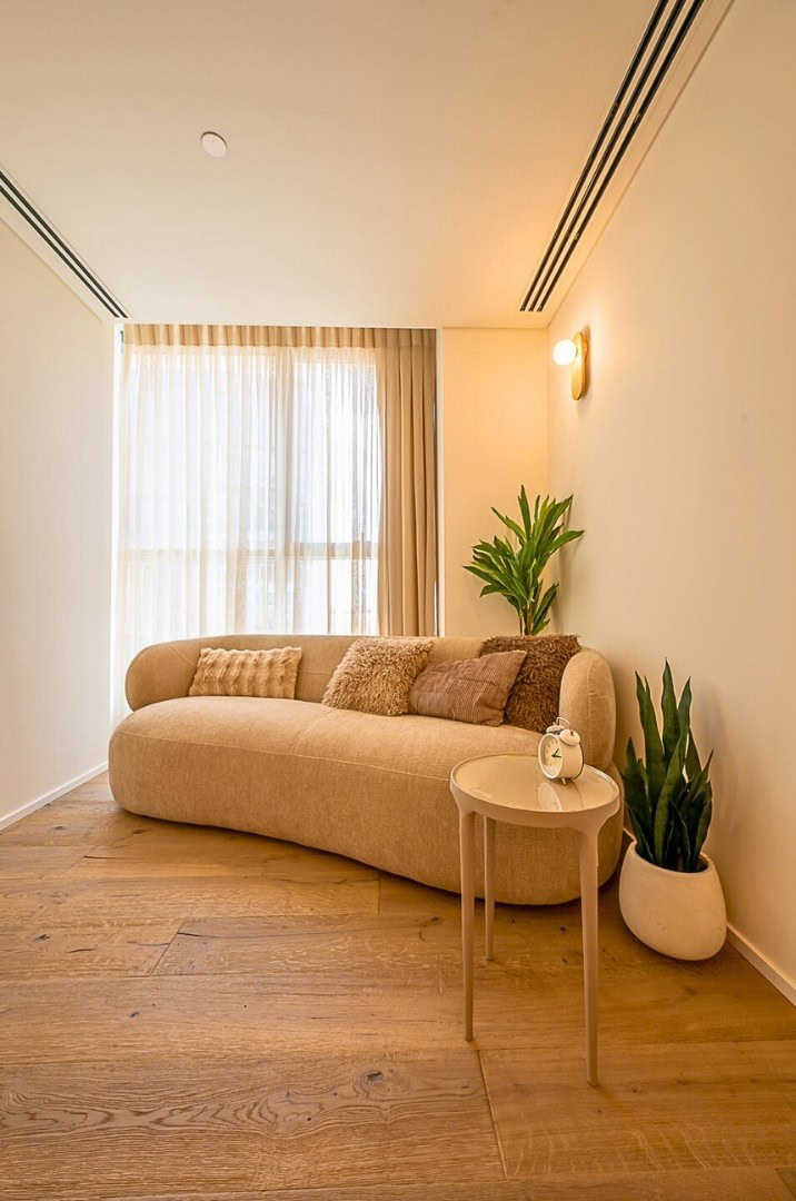 סלון נעים ומודרני עם ספה מעוגלת בצבע בז', כריות רכות, שולחן צד עגול קטן עם קישוט קרמיקה, שני עציצים ירוקים, ריצוף עץ ווילונות שקופים המאפשרים כניסת אור טבעי.