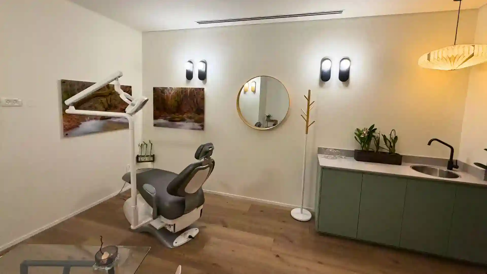 מרפאת שיניים מודרנית כוללת כיסא רופא שיניים מדגם Satori Shibui, תמונות קיר בהשראת הטבע, מראה עגולה, תאורה מינימליסטית, מתלה מעילים, צמחים, כיור וארונות בצבע ירוק על גבי ריצפת עץ. החלל מואר ומסודר.