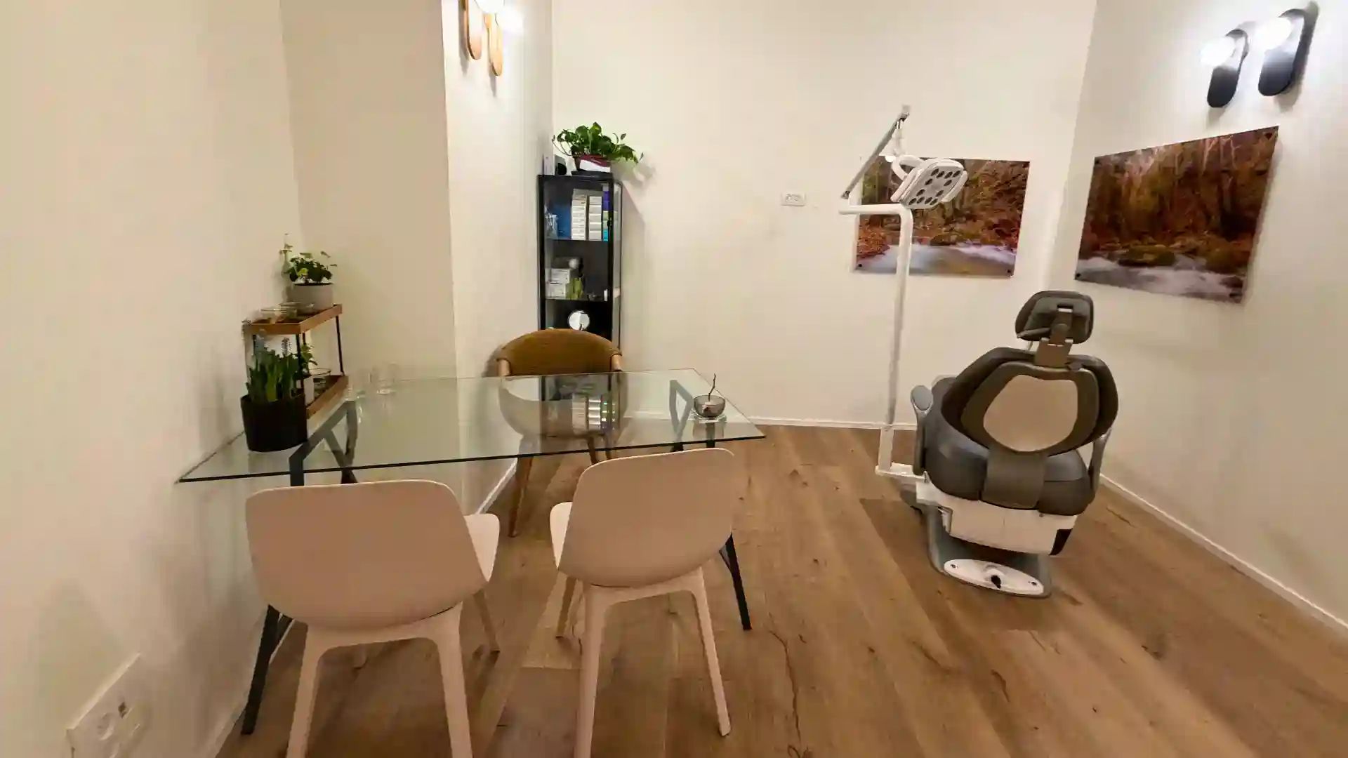 מרפאת שיניים נקייה ומודרנית כוללת שולחן זכוכית בהשראת סגנון "סאטורי שיבוי" עם שני כיסאות, כיסא לבדיקת שיניים, מדף ספרים שחור, עציצים ותמונות נוף על גבי רצפת עץ בהירה.