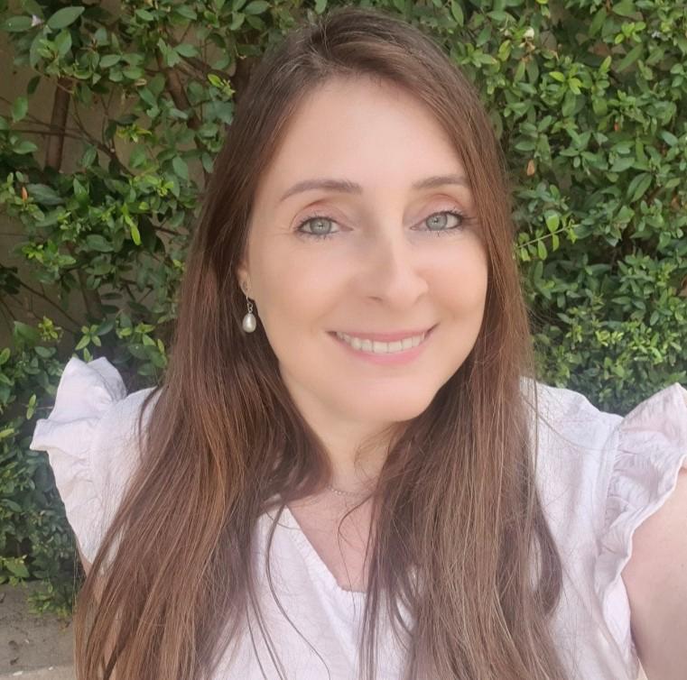 אישה עם שיער חום ארוך וחולצה ורודה בהירה מחייכת על רקע ירוק ועשיר בעלים.